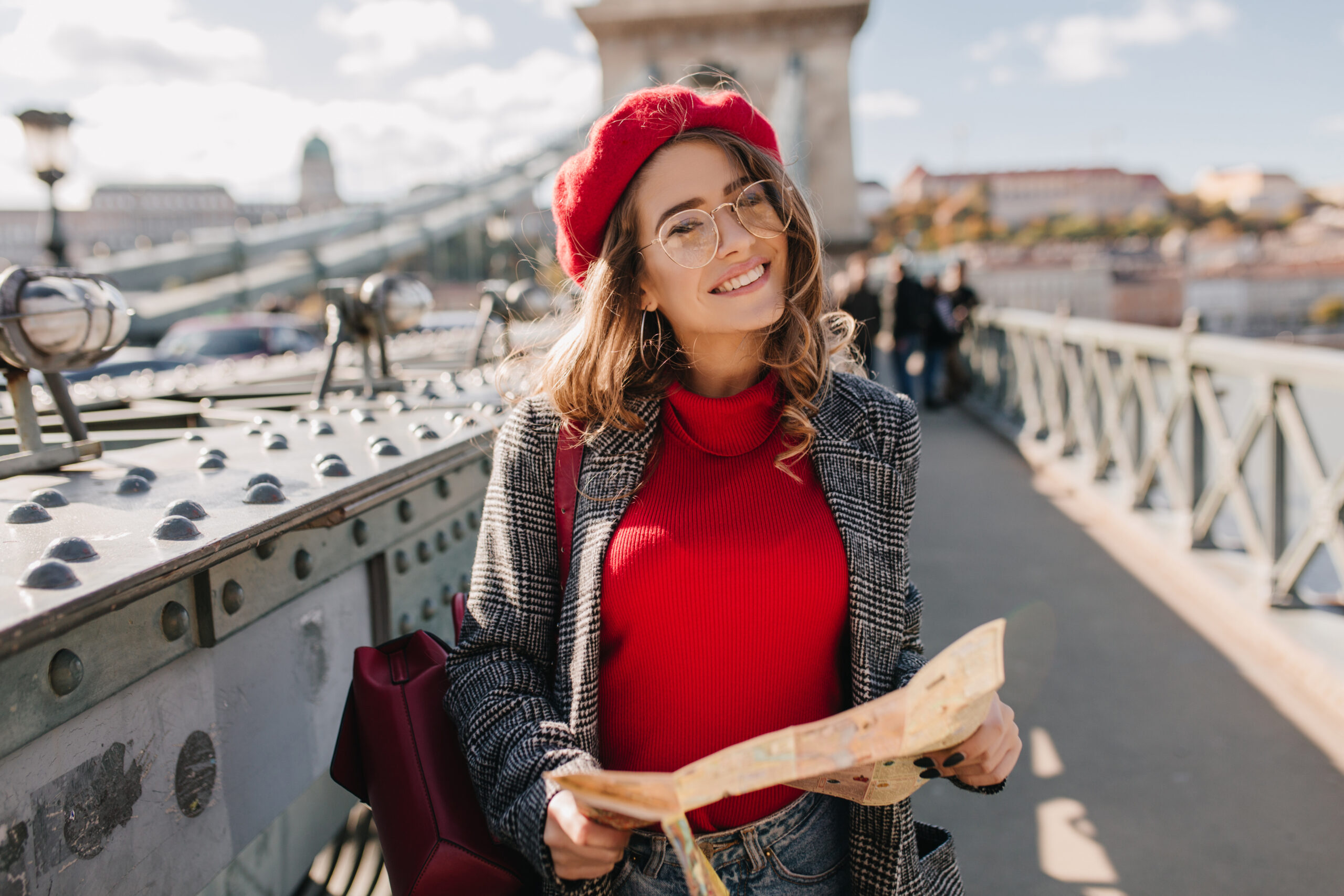 Pleased brunette woman with red backpack posing on bridge on urban background. Attractive white girl in glasses using map, exploring european city.