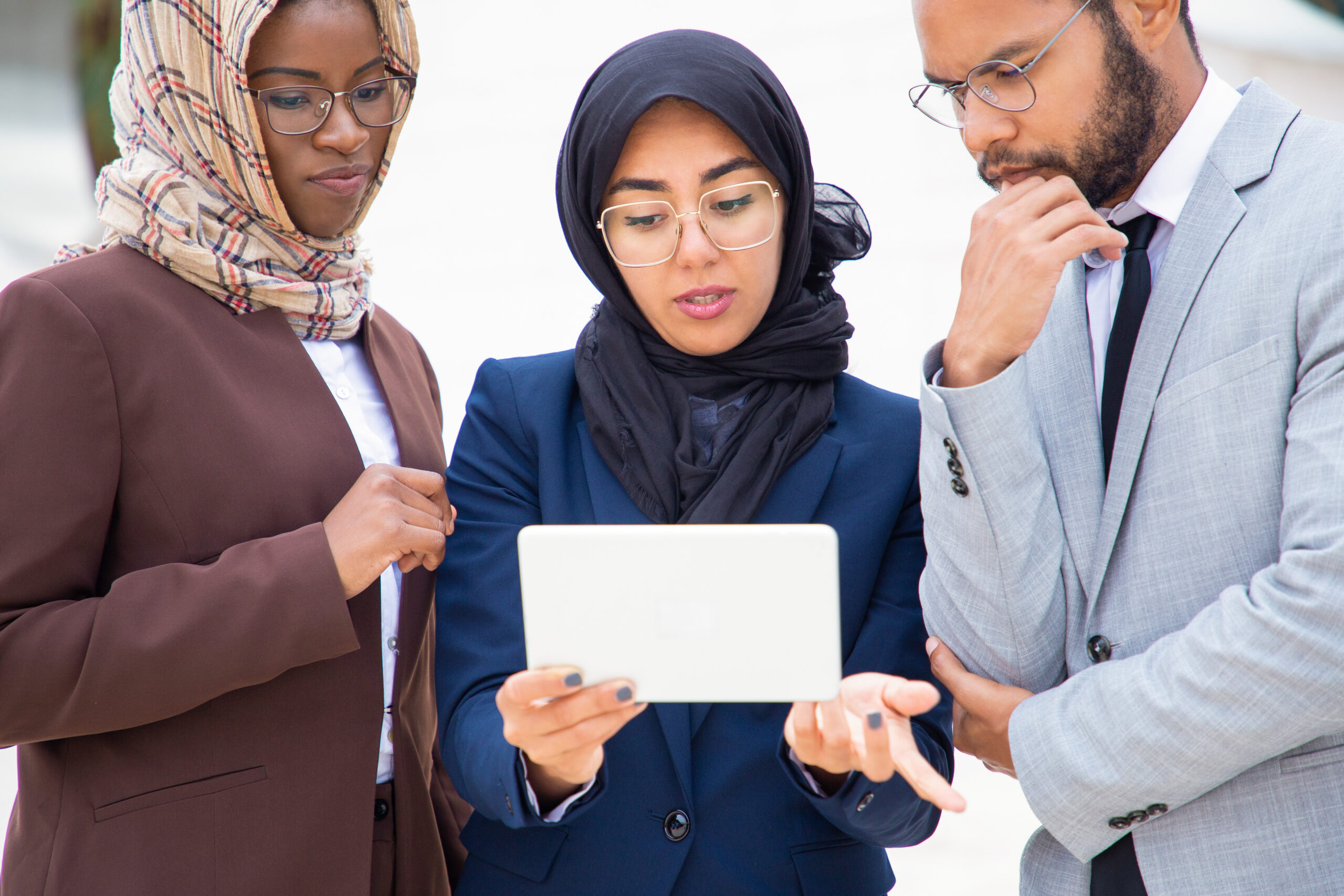 Serious diverse business group watching and discussing content on tablet outside. Businessman and Muslim businesswomen standing outdoors and using tablet together. Digital communication concept